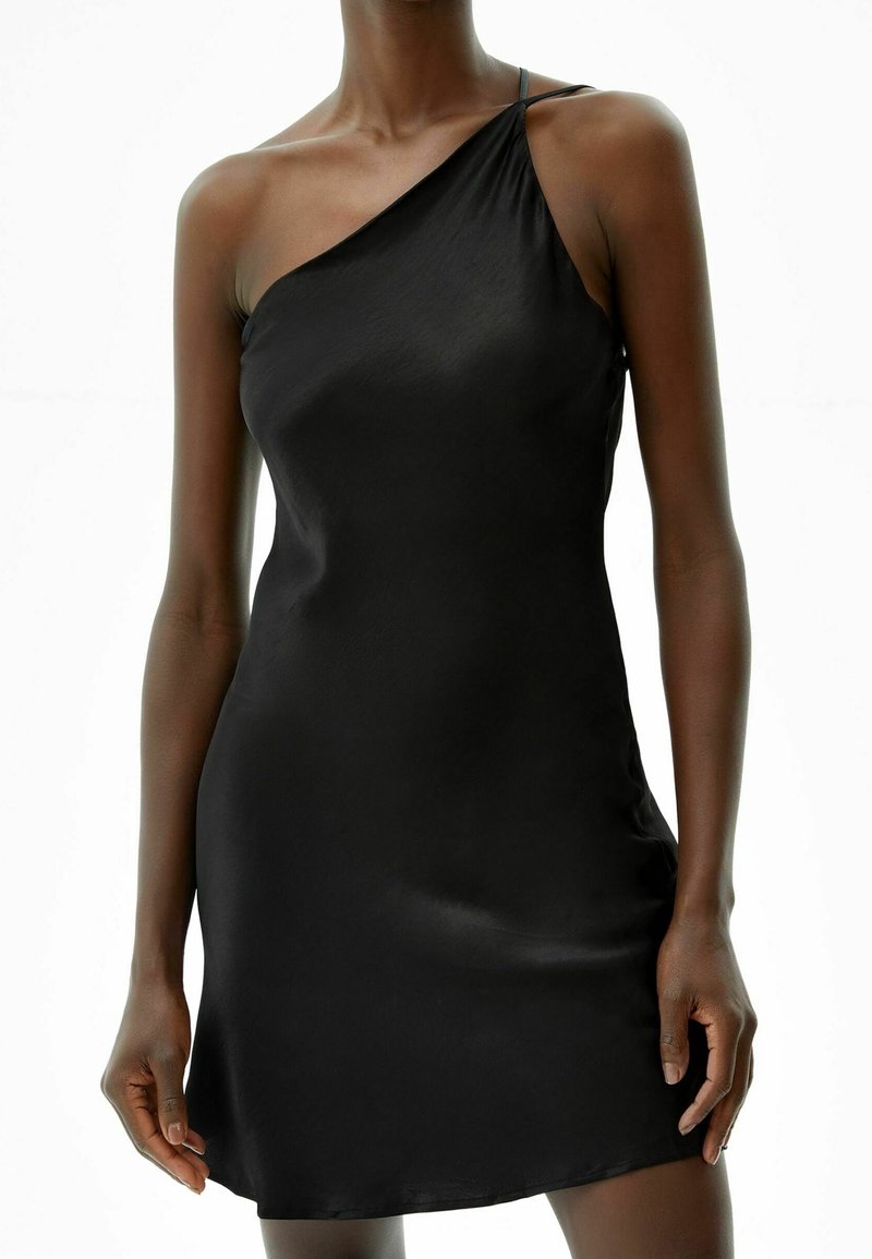 Femme portant une mini-robe noire en satin sans manches avec une fine bretelle sur l'épaule gauche et un décolleté diagonal.