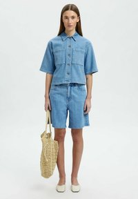Giacca di jeans azzurro chiaro con maniche corte e due tasche frontali, abbinata a pantaloni corti in denim dello stesso colore. Il modello tiene in mano una borsa intrecciata beige e indossa scarpe chiare.