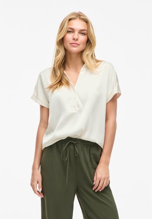 Plavolaska ženska z oblečeno kratko kremasto bluzo s kratkimi rokavi in olivno zelenimi hlačami z vrvico, stoji pred enobarvnim svetlim ozadjem.