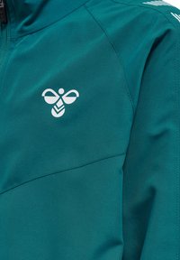 Veste teal avec un design zippé, en tissu léger, et un logo blanc sur la poitrine, avec des détails de coutures visibles et une texture lisse.