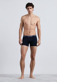 Cuecas boxer pretas de algodão com uma cintura contrastante apresentando o logótipo da Calvin Klein, usadas por um modelo em pé sobre uma superfície cinzenta.