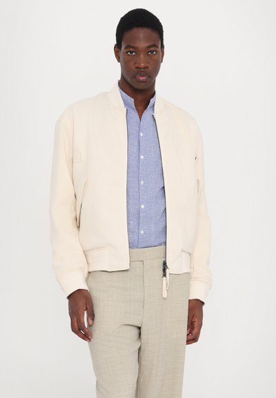 Veste bomber beige clair avec des poignets côtelés, fermeture éclair et poches latérales, portée sur une chemise bouton bleu et un pantalon beige texturé.