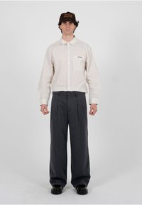 Homme portant une chemise à rayures boutonnée rentrée dans un pantalon gris à jambes larges, des chaussures noires et une casquette marron avec le texte "L'art d'être en retard".