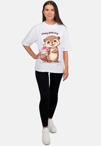Wit oversized t-shirt met een cartoonbeeld van een otter die een verjaardagstaart vasthoudt. Gecombineerd met zwarte leggings en witte sneakers.