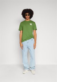 Žalias medvilninis T-shirt su baltu logotipu, derinamas su šviesiai mėlynais džinsais ir baltais sportbačiais. Modelis dėvi tamsius akinius nuo saulės. Paprasta fono.
