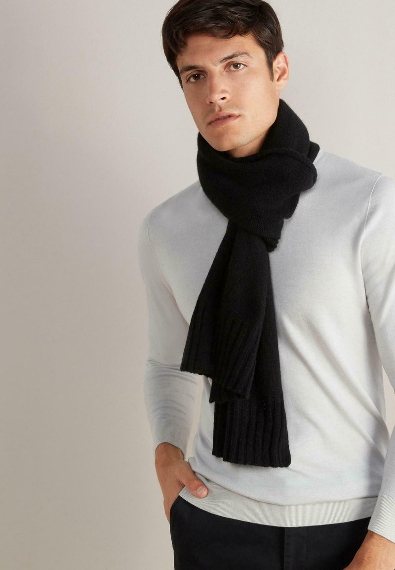 Sort strikket tørklæde med ribbet tekstur, draperet omkring halsen, båret over en lysegrå sweater. Tydelig farvekontrast med en glat finish.