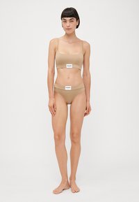 Beige kaheosaline komplekt, mis sisaldab lühikeste õlgadega crop-topi ja sobivaid kõrge vöökohaga aluspükse, mõlemal on logo sildid. Sile kangateksuur.