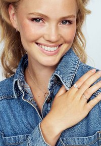 Jeansjacka med blå nyans, prydd med metallknappar. Modellen bär silverörhängen, ett tunt halsband och två ringar med dekorationer.