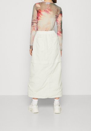 Kobieta ubrana w długą białą spódnicę, kolorową bluzkę z długim rękawem tie-dye, białe skarpetki i masywne białe sneakersy, zwrócona tyłem na jednolitym tle.