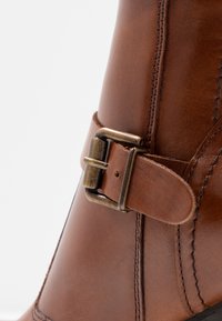 Bottine en cuir marron à la forme incurvée, avec une boucle en métal bronze et des coutures apparentes sur le côté. Texture lisse.