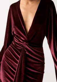 Robe en velours marron avec manches longues, décolleté en V profond et détail torsadé sur le devant. Caractérisée par une texture froncée et un tissu lisse.