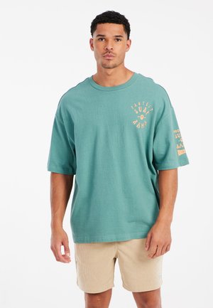 Miętowy bawełniany t-shirt o luźnym kroju, z graficznym nadrukiem w pomarańczowym kolorze na lewej piersi i rękawie. Nosi się go z beżowymi szortami welurowymi.