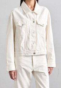 Bílá riflová bunda s límcem, zapínáním na knoflíky a dvěma náprsními kapsami. Má krátký střih a viditelné detaily švů.