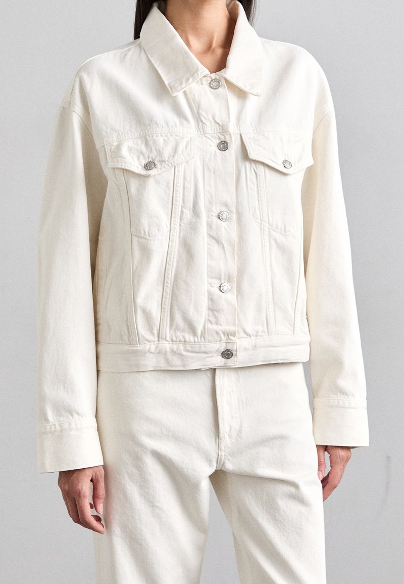 Bílá riflová bunda s límcem, zapínáním na knoflíky a dvěma náprsními kapsami. Má krátký střih a viditelné detaily švů.