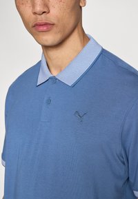 Μπλε πόλο πουκάμισο με υφασμένη ανοιχτό μπλε γιακά, δύο κουμπιά και σκούρο μπλε κεντημένο λογότυπο στο αριστερό στήθος. Ομαλή υφή βαμβακιού.