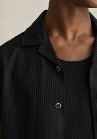 Cămașă neagră cu mânecă scurtă, textură ribbed, închidere cu nasturi și guler cu capse. Purtați-o peste un maiou negru asortat.