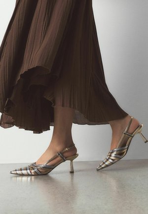 Beine in einem wehenden braunen Rock, die einen Schritt nach vorne machen und metallisch spitze Slingback-Pumps mit niedrigen Stilettoabsätzen auf einem glatten Boden tragen.