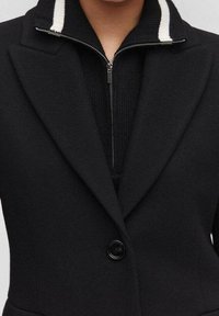 Zwarte blazer met een gestructureerd ontwerp, voorzien van een geknipt kraag en een zilveren rits. Witte streepdetail bij de kraag, sluiting met één knoop.