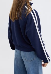 Sudadera azul marino oversize con rayas blancas en las mangas, cuello alto y ribete acanalado. Combinada con vaqueros de talle alto en azul claro.