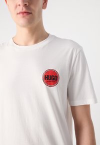 Valge puuvillane T-särk, millel on ümmargune punane plaastriga, kus on musta värviga kiri "HUGO" ja "WINNING BLEND", mille ääres on ruuduline muster.