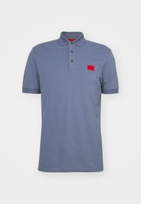 Pikétröja i ljusblått bomull, med en röd logotypapplikation, tvåknapppolk och ribbade ärmslut. Slät textur, avslappnad design.