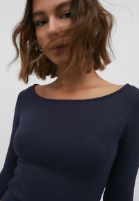 Haut long à manches avec côtes de couleur bleu marine, décolleté large, silhouette ajustée et texture douce. Le tissu semble lisse et extensible.