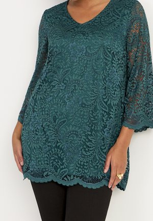 Woman wearing teal lace blouse with floral pattern and scalloped edges, paired with black pants, hands resting by sides.