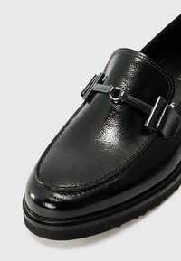 Mocassin en cuir verni noir avec un bout rond, arborant une finition texturée, du matériel décoratif noir et une semelle en caoutchouc robuste.
