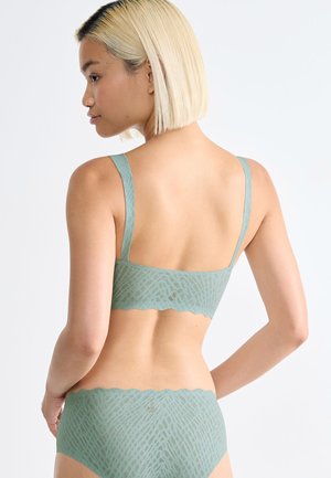 Mujer con cabello rubio corto que lleva un conjunto coordinado de sujetador y ropa interior de estampado verde claro, vista desde atrás sobre un fondo blanco liso.