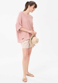 Lichtroze oversized gebreide trui met brede mouwen, gecombineerd met beige shorts en sandalen. Houdt een beige hoed vast, minimalistisch ontwerp.