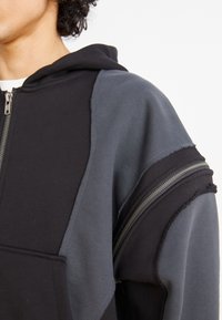 Black and gray hooded sweatshirt with a zipper, featuring color-blocked panels and frayed edges. Fabric appears soft and textured.