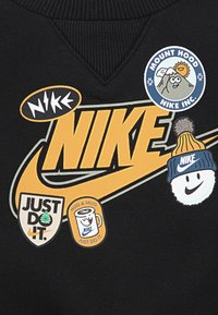 Crna dukserica s velikim žutim logotipom "NIKE" i raznim šarenim zakrpama, uključujući "JUST DO IT" i razigrani dizajn planine.