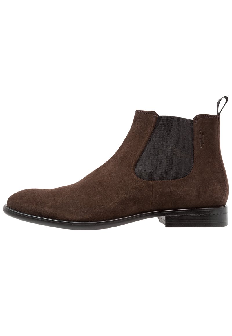 Bota Chelsea de ante marrón con paneles laterales elásticos negros, punta redondeada lisa y un tacón bajo apilado. Cuenta con una lengüeta en la parte posterior.