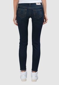 Donkere denim skinny jeans met een mid-rise taille, voorzien van opvallende achterzakken met stikdetails en een gladde textuur. Draagt witte sneakers.