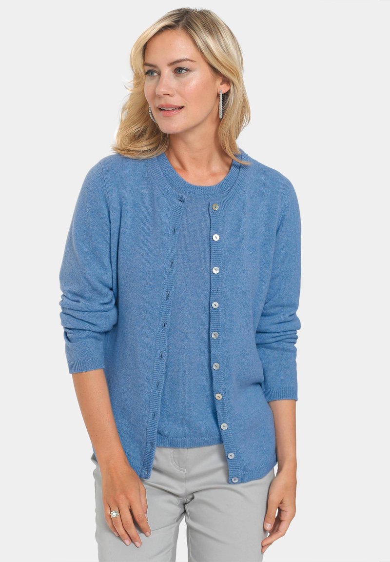 Blauer Cardigan aus weichem Material, mit Rundhalsausschnitt, Knöpfen vorne, gerippten Bündchen und lockerer Passform. Kombiniert mit einem hellblauen Oberteil.