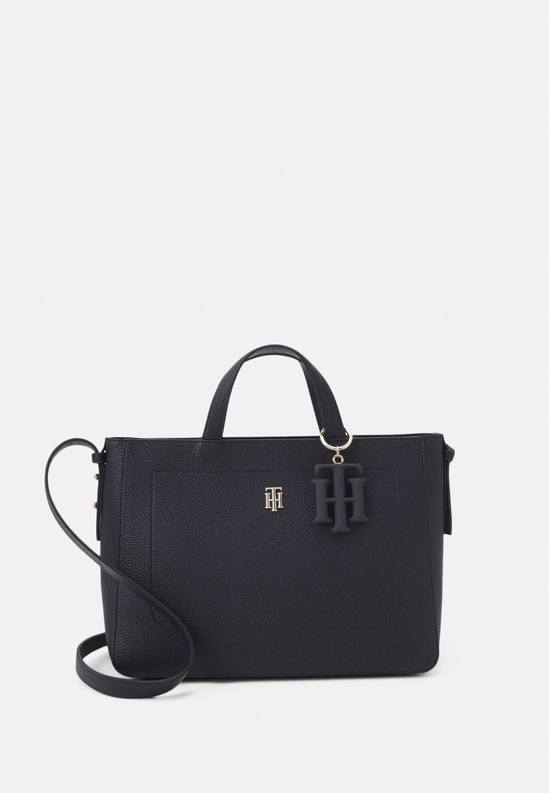 Sac à main en cuir noir avec poignées courtes, longue bandoulière, petit logo doré et grand pendentif noir avec initiales suspendues.