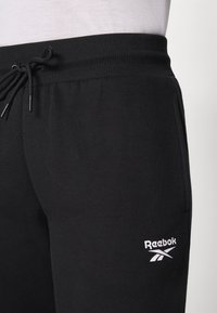 Črne trenerke iz mehkega materiala, z rebrastim pasom in vrvico za zategovanje. Vključujejo majhen bel detajl logotipa Reebok.