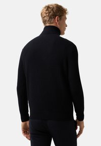 Blonder Mann trägt einen schwarzen, gerippten Rollkragenpullover und schwarze Hose und ist von hinten vor einem schlichten weißen Hintergrund zu sehen.