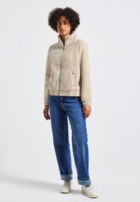 Giacca in pile beige con colletto alto, chiusura con zip e polsini elastici. Abbinata a jeans blu arrotolati e sneakers bianche.