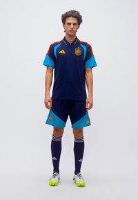Young man standing straight wearing dark blue and light blue soccer kit with red and yellow stripes, logos on chest and shorts, and neon yellow cleats.