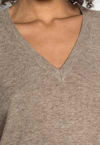 Maglione beige a V con un motivo di maglia testurizzata. Il tessuto è morbido e leggero, con una leggera lucentezza e dettagli a coste sul collo.