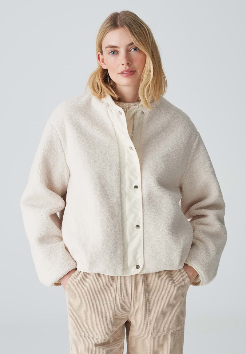 Veste de couleur crème, texturée, avec un extérieur doux et duveteux et une doublure intérieure lisse contrastante. Dotée d'une fermeture à boutons-pression et d'une coupe décontractée.