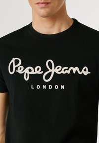 Homme portant un t-shirt noir avec le logo blanc "Pepe Jeans London" sur la poitrine, fond uni.