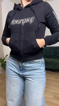 Pessoa vestindo um casaco com capuz preto com fecho e texto "wasted" e jeans azul claro, mãos nos bolsos do casaco, em pé dentro de casa perto de uma planta e de um sofá.