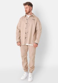 Beige katoenen utilityjack met drukknoopsluiting en logo op de borst, gecombineerd met bijpassende tapered broek en witte sneakers. Soepele textuur, relaxte pasvorm.