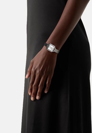 Brazo de piel oscura que lleva un reloj de pulsera de cara cuadrada y plateado con una correa de metal, apoyado contra un vestido negro.