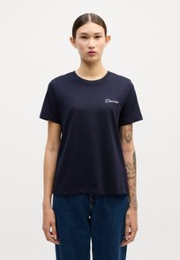 Mörkblå bomull T-shirt med korta ärmar, rund halsringning och en liten vit broderad logotyp på vänster bröst. Bärs tillsammans med högmidjade jeans.