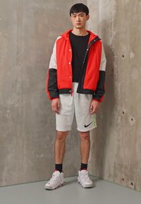 Giacca a vento rossa e nera con maniche bianche, t-shirt nera, pantaloni corti bianchi con logo Nike e scarpe da ginnastica bianche con dettagli rossi.