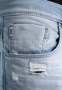Ljust blå denimjeans med en slitna design, inklusive en riven ficka och metallnitar. Slen yta med synliga sömdetaljer.