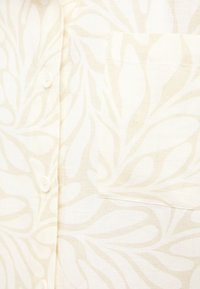 Light-colored linen shirt with a subtle leaf pattern, featuring a patch pocket and buttons along the front. Soft texture and breathable fabric.